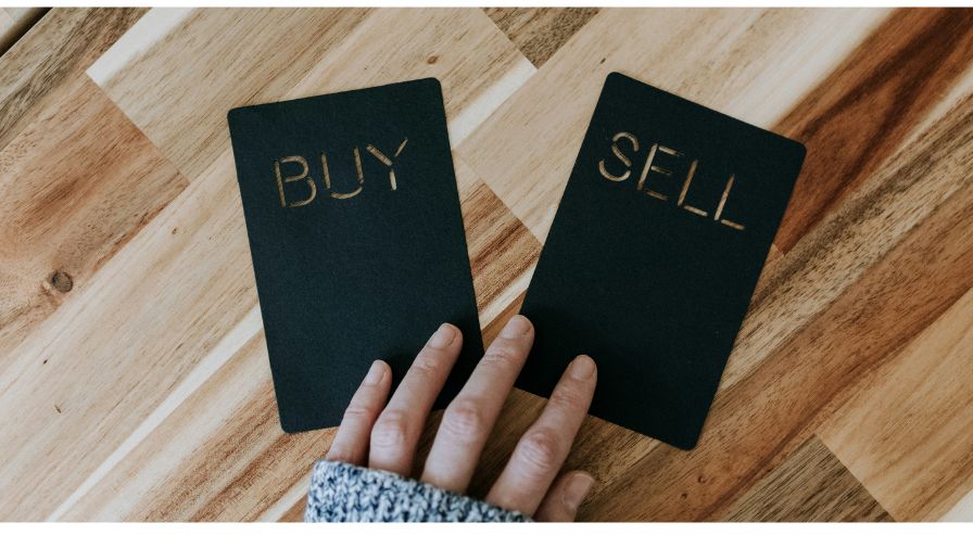 Hand reaching towards black cards labeled 'BUY' and 'SELL' on a wooden surface, representing the concept of buying and selling in financial decisions.