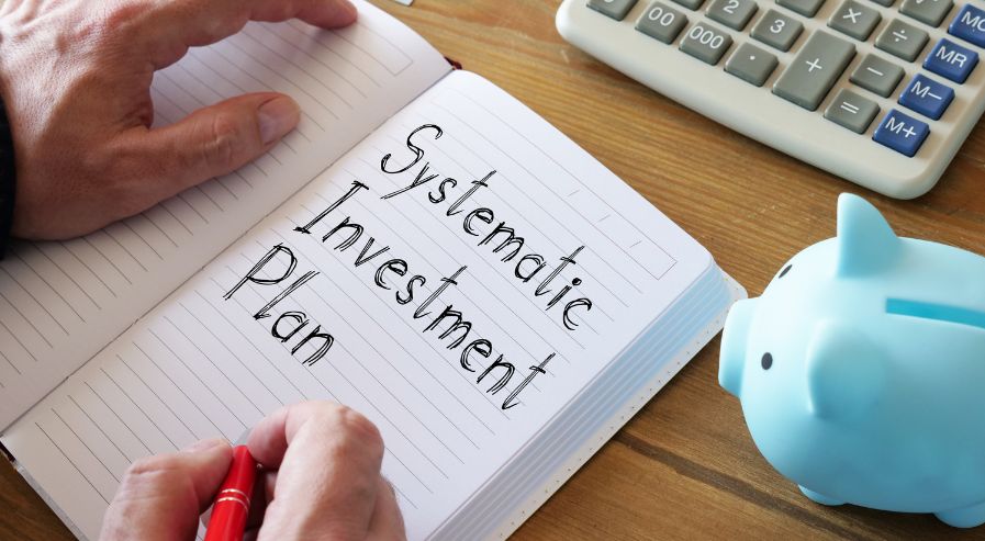 Notebook with 'Systematic Investment Plan' written on it, held by a person with a red pen, next to a blue piggy bank and a calculator.